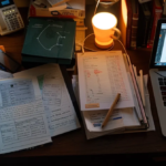 a person sitting at a desk with a laptop, notebook, and pen, surrounded by study materials and charts, preparing for their cmhc mli select application.